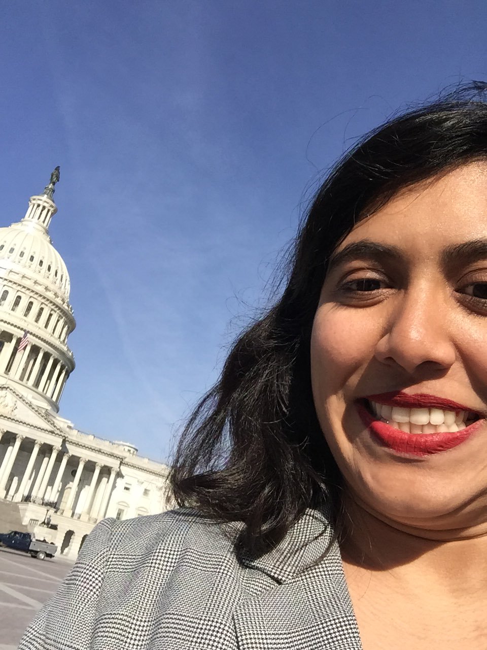 A selfie of an Indian woman in front of the US Capitol. She is has her hair down and is wearing a black and white checked blazer with red lipstick.