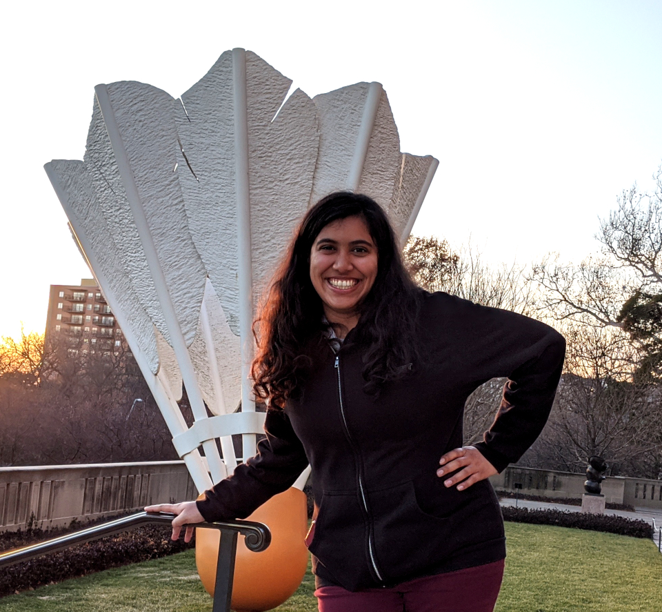 A young Indian woman in a black jacket and maroon pants with her hair down poses with one hand on her hip in front of a giant badminton shuttle on a lawn at sunset. The background has trees missing their leaves and a short brick apartment building.